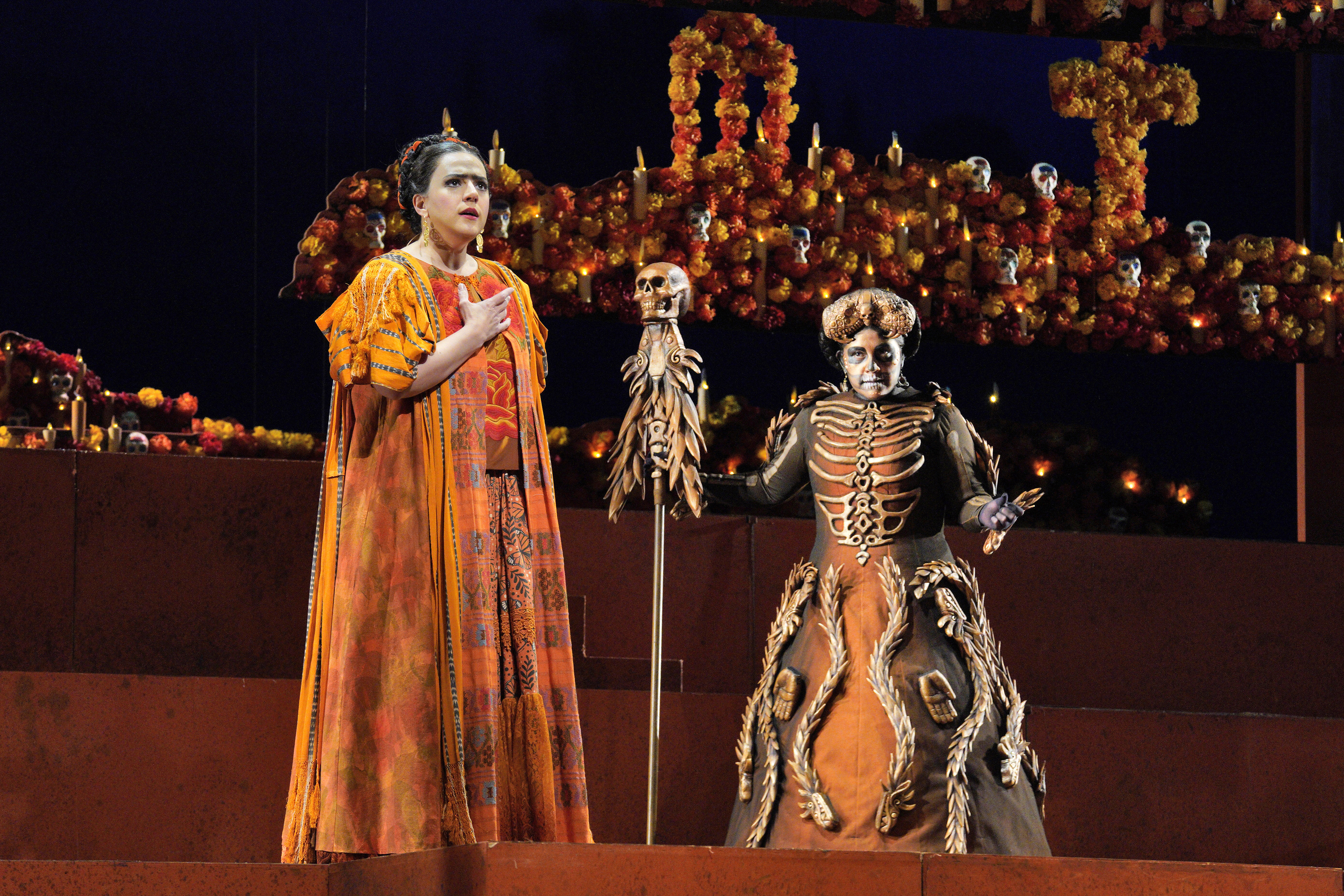Scene from Frida Two women in costumes standing on a stage in front of skeletons.