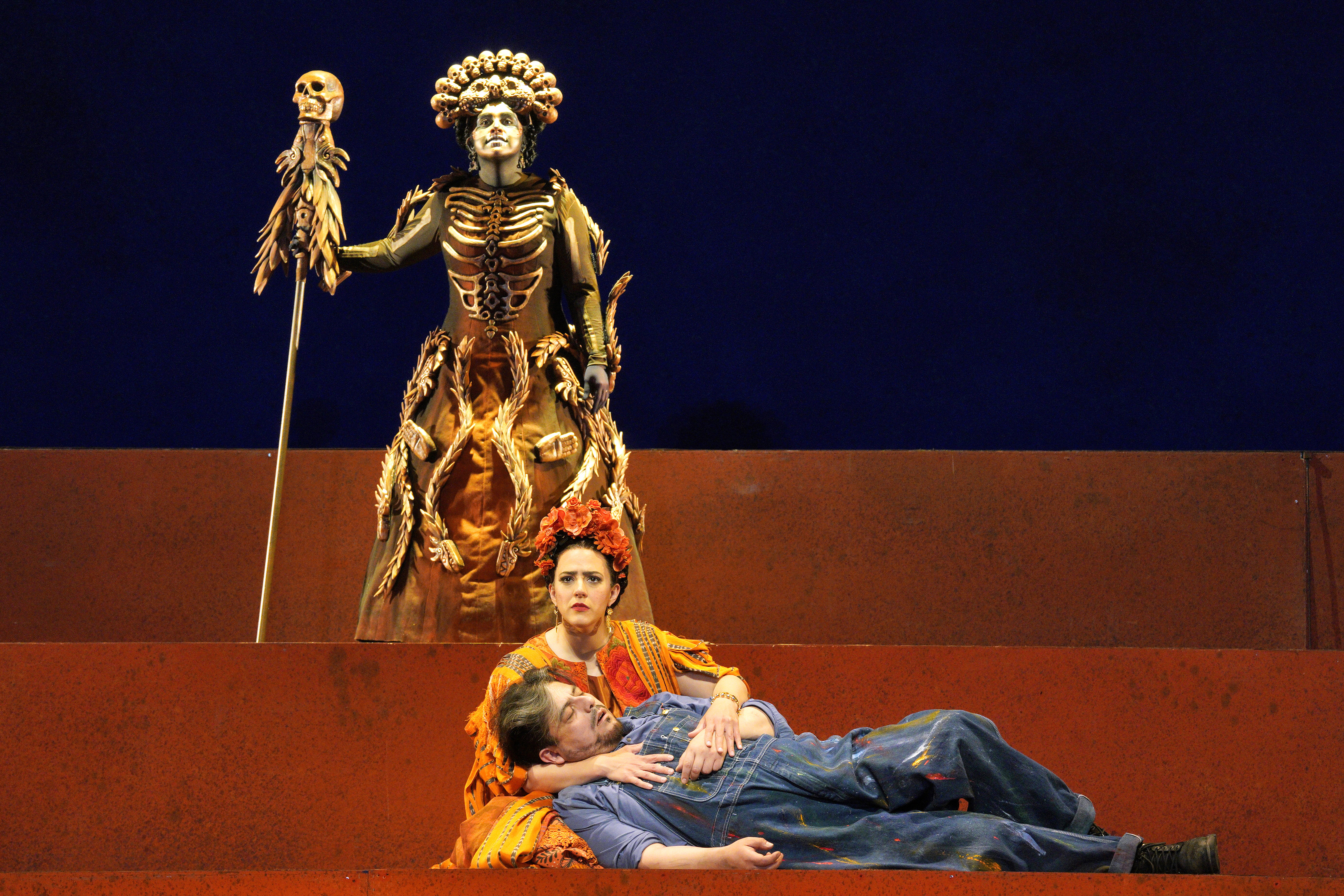 Scene from Frida of a woman holding a man laying on the floor while a person in a skeleton costume stands behind them.