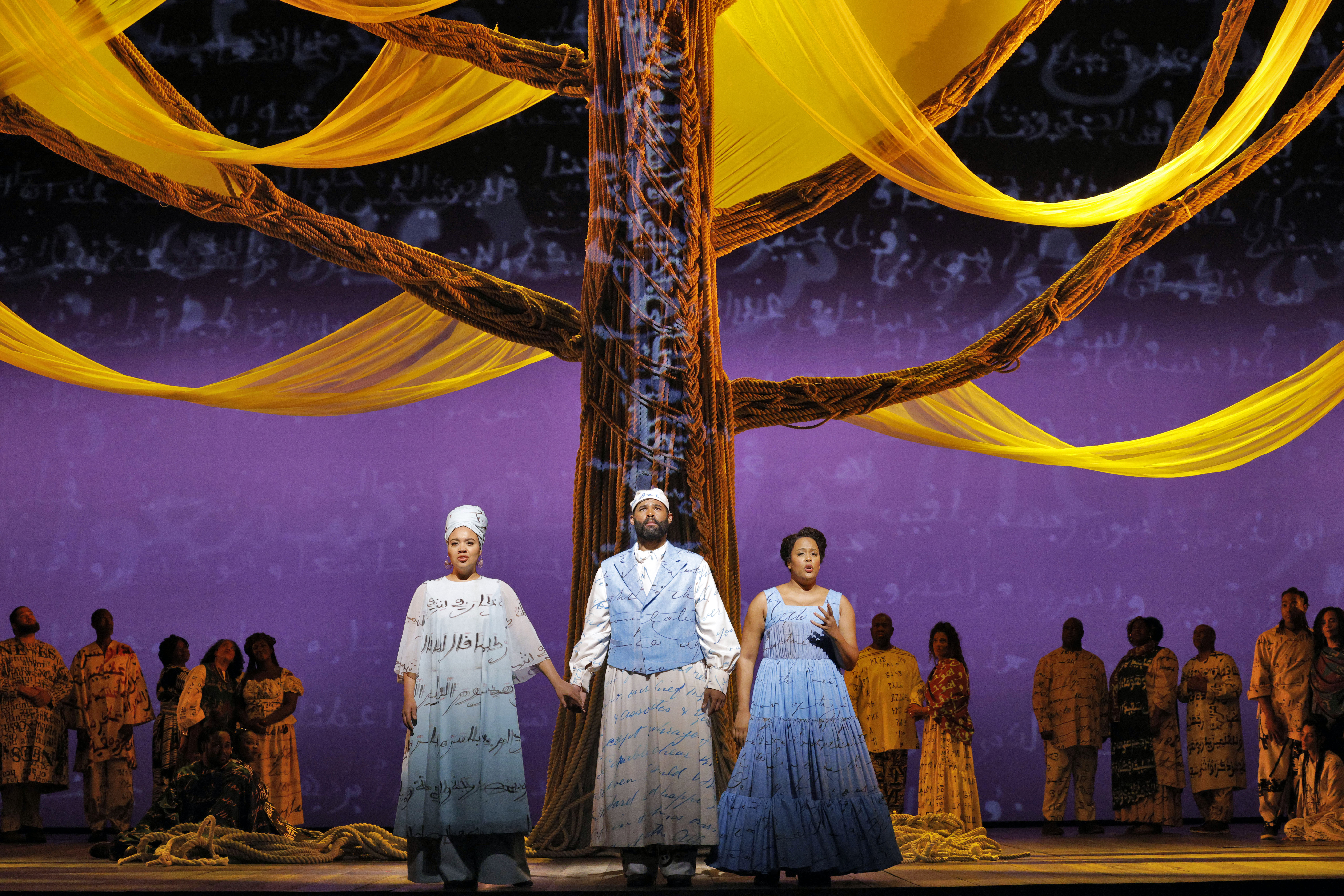 three people standing in front of tree with groups in the background. two (a man and woman) are holding hands